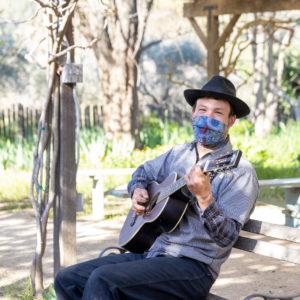A man wearing a black hat while strumming a guitar and sitting on a bench.