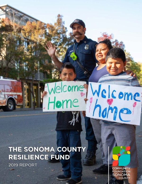 A Latina mother standing behind her two children who are holding signs reading "welcome home" and are standing with a Healdsburg firefighter in downtown Healdsburg. They are welcoming home people evacuated during the Kincade Wildfire.