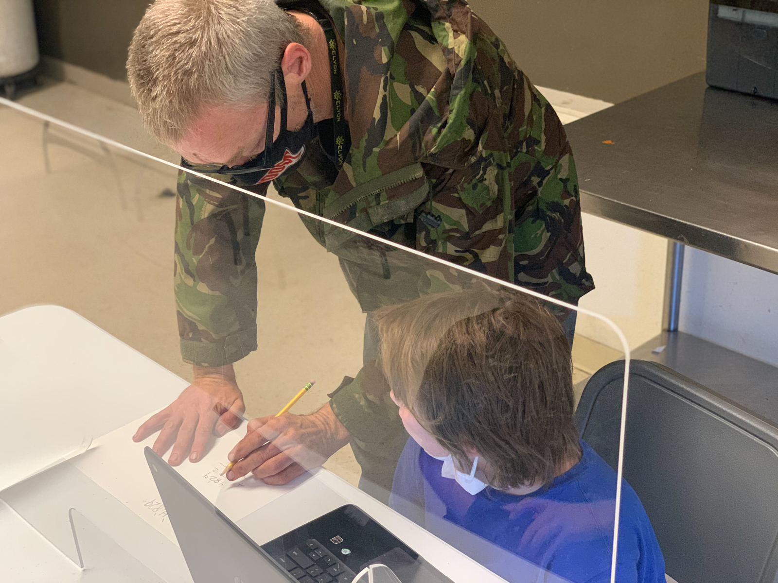 A man dressed in Army camouflage helping a young boy with his school work.