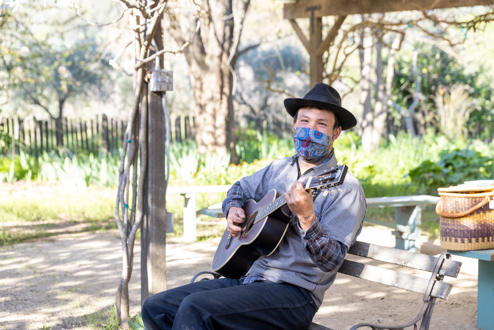 A man wearing a black hat while strumming a guitar and sitting on a bench.