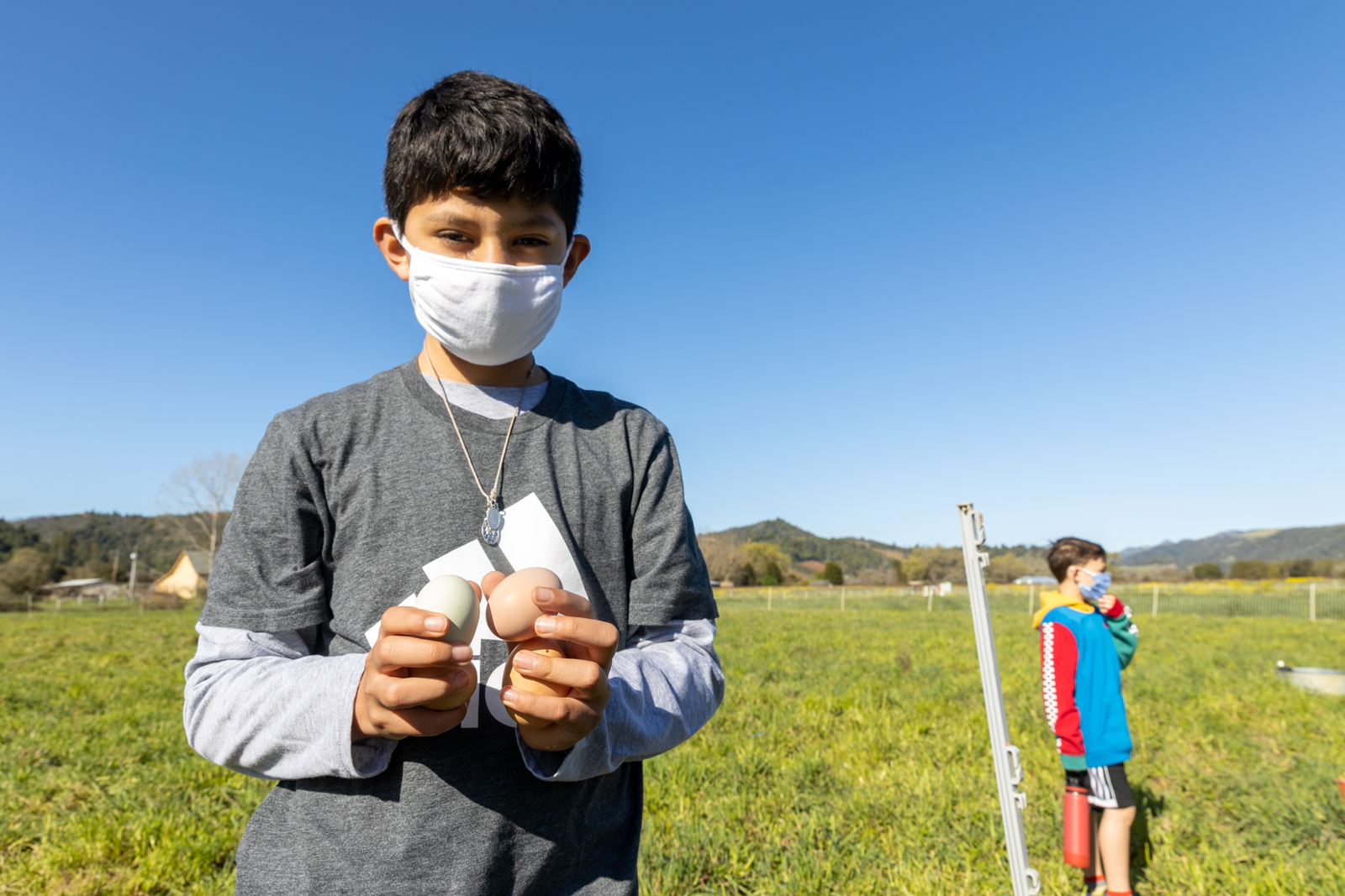 A boy on a farm wearing a white mask holding a chicken egg in each hand.