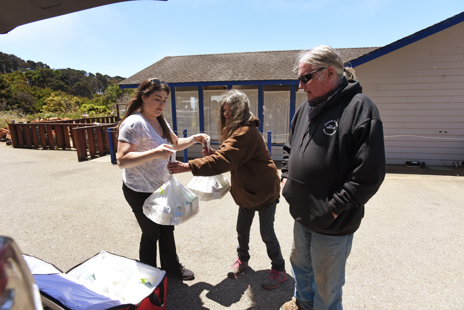 Elder community members picking up his food from a young lady holding bags of food.