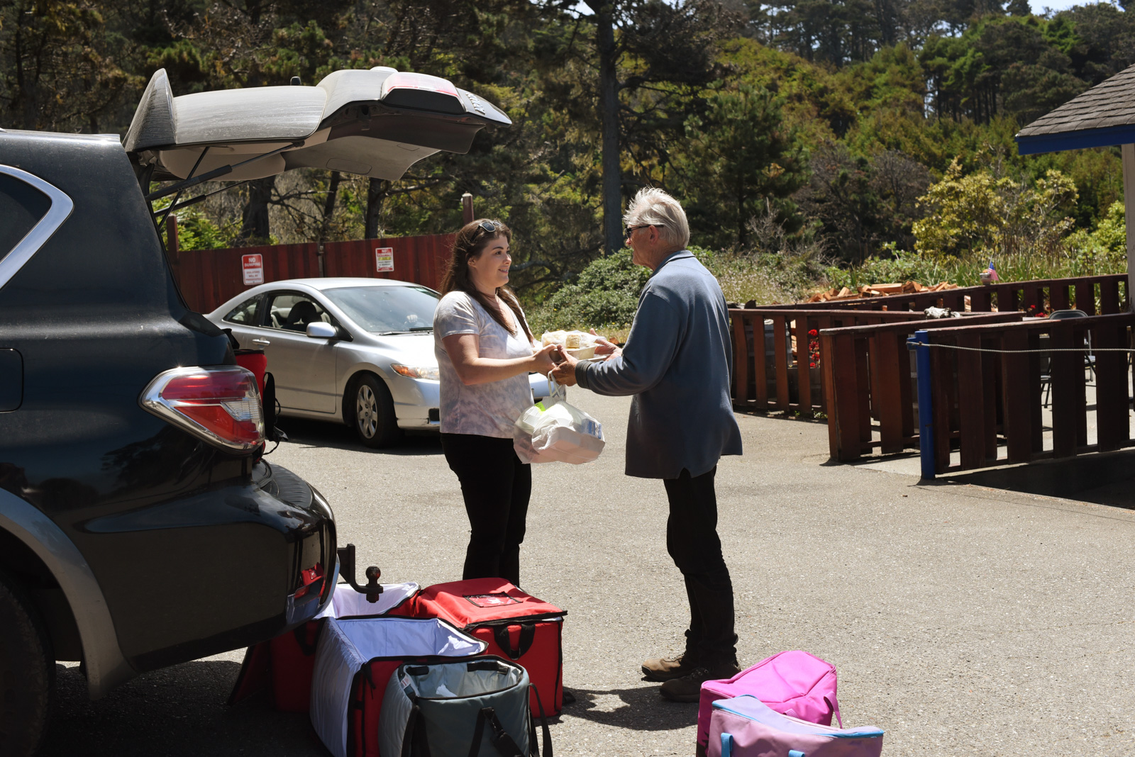 An elder community member picking up his food from a young lady holding trays and bags of food.