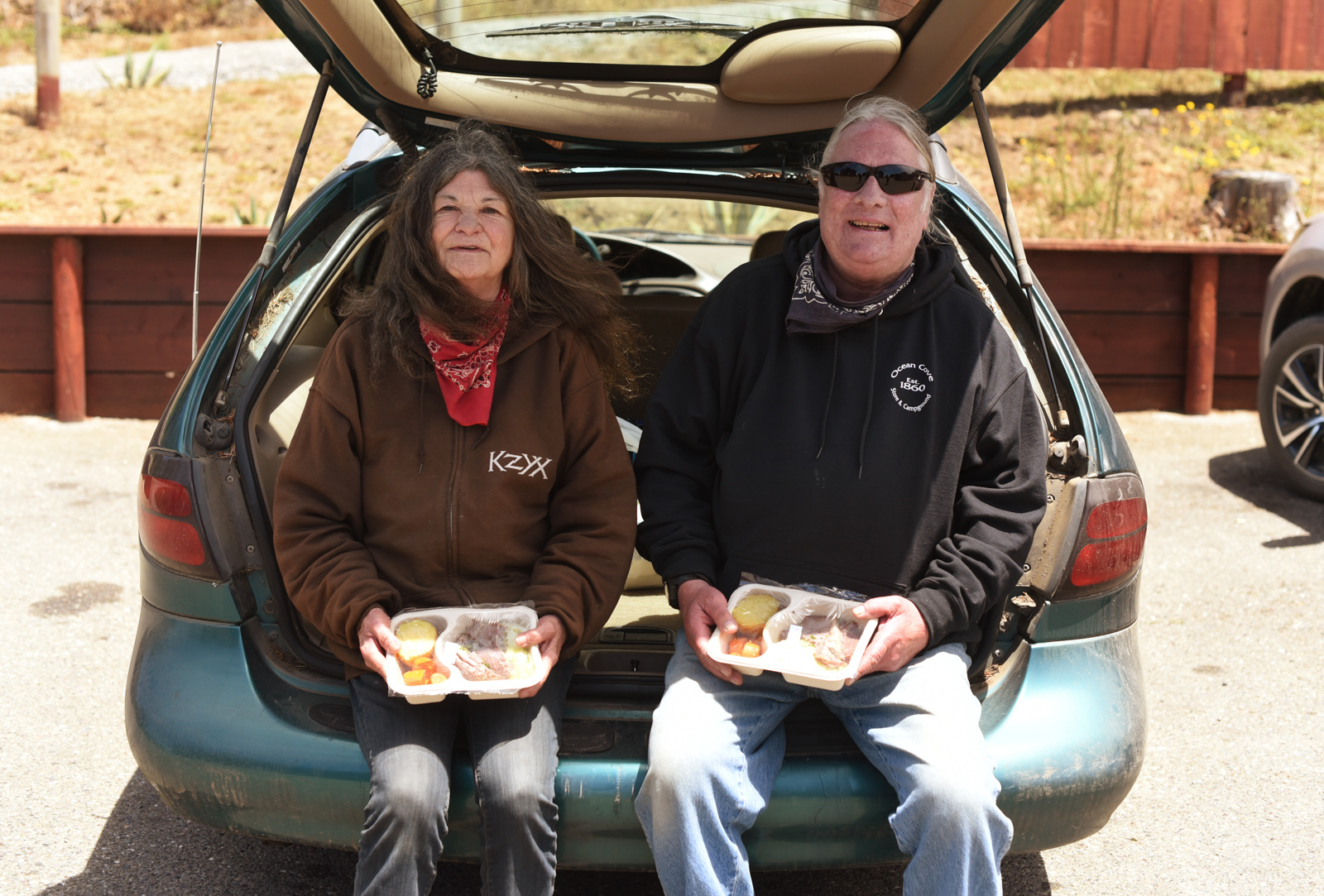 Elder community members holding food trays as they sit on the trunk of their car.