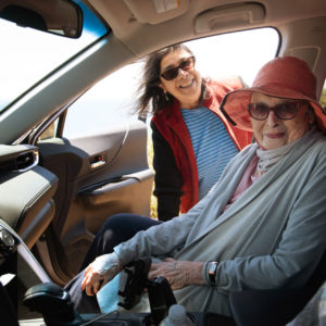 An older woman sits passenger side in a car as her friend stands outside next to her.