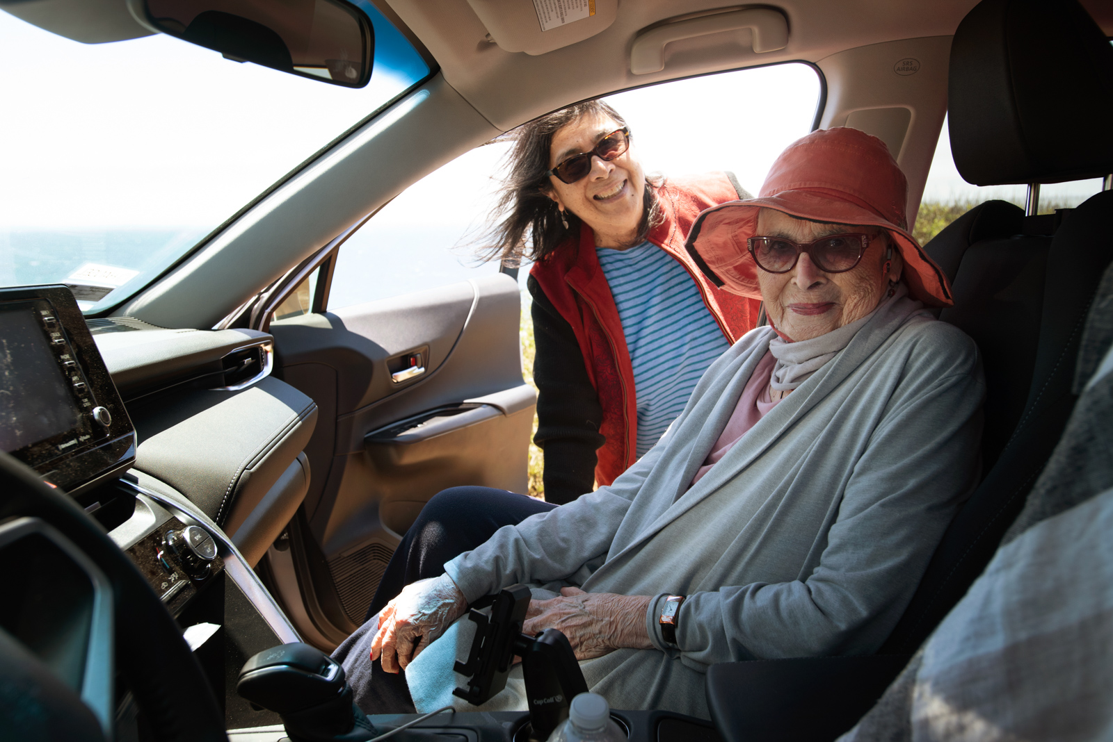 An older woman sits passenger side in a car as her friend stands outside next to her.