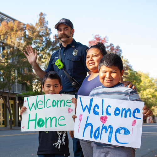 A Latina mother and two children hold signs reading "welcom home" and are standing with a Healdsburg firefighter in downtown Healdsburg. They are welcoming home people evacuated during the Kincade Wildfire.