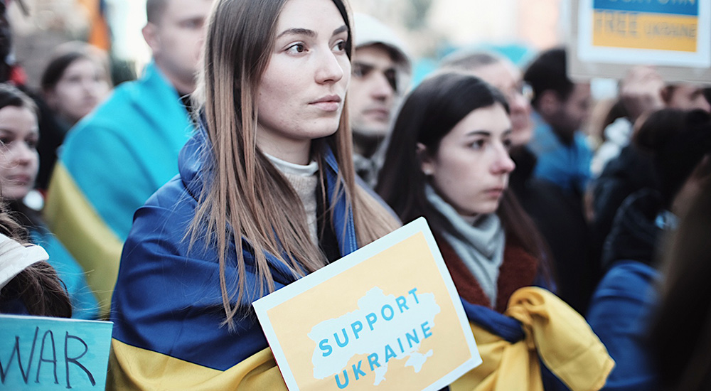A young gift draped in a Ukrainian flag holds a sign reading "support Ukraine" at a peace rally in Brussels, Belguim. Phoe taken by Bartosz Brzezinski on flickr https://www.flickr.com/photos/bartosz/51908400925/