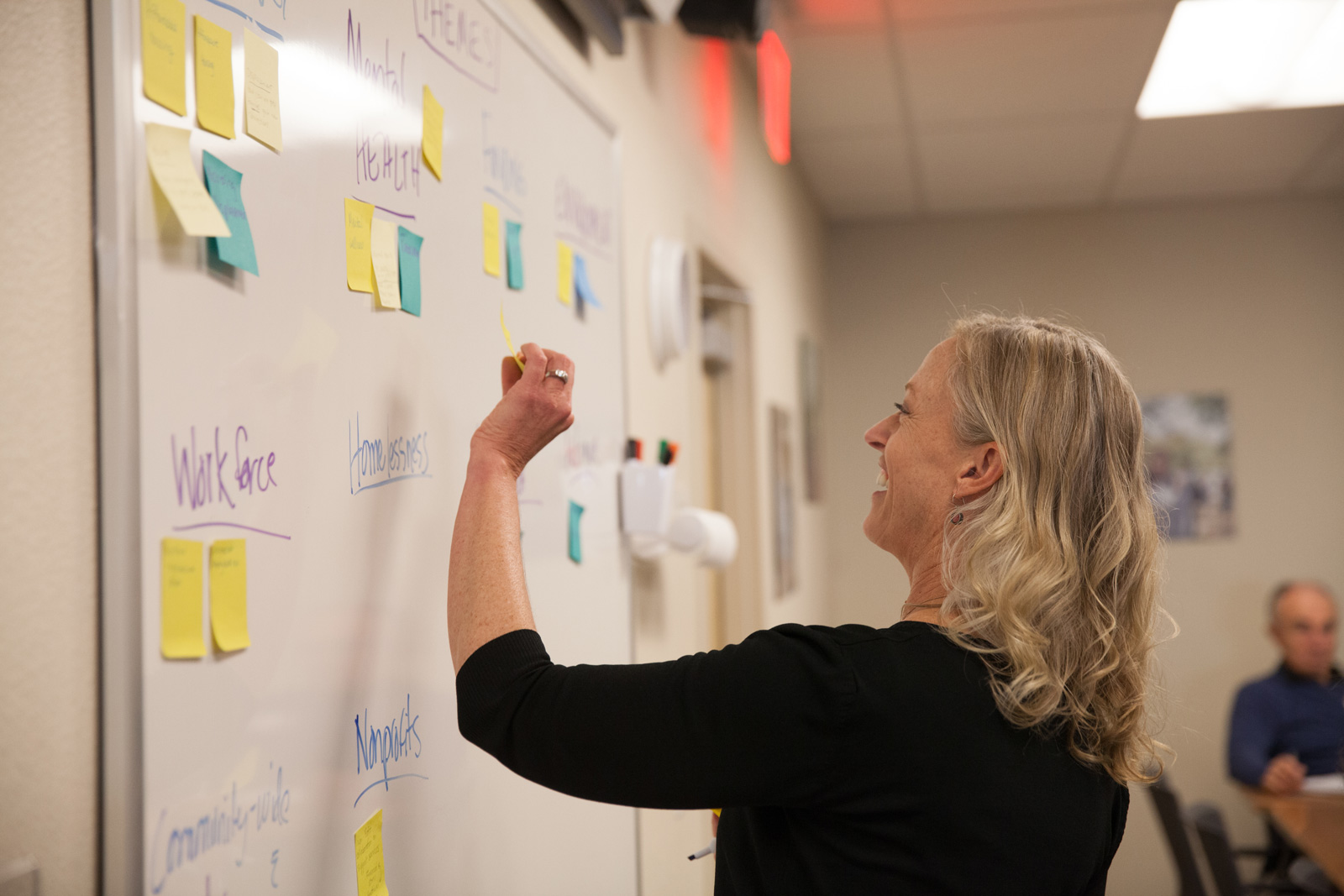 Karin Demarest stands in front of a white board covered in sticky notes, she is arranging them into categories, including workforce, mental health, homelessness, nonprofits.