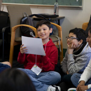 Elementary school age youth sitting in a circle in a classroom, one is holding up a piece of paper, the others are smiling and listening.