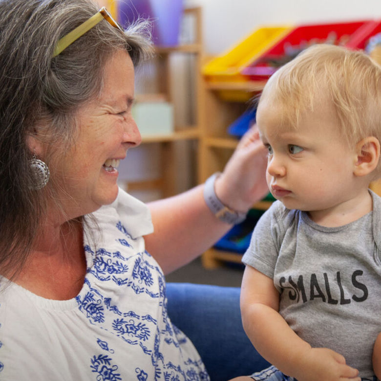 A woman smiling at a young child in a daycare. Text: Julia L. Grant Basic Human Needs Grants Program