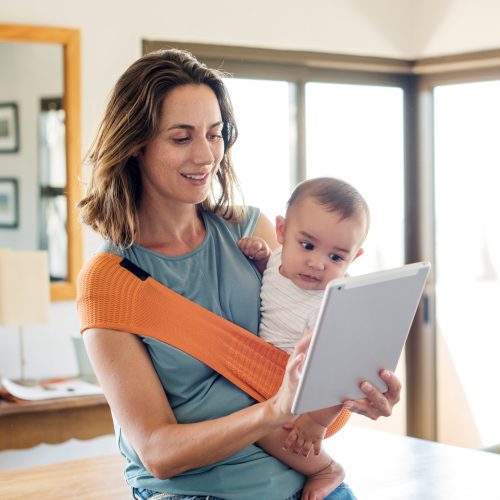 Mother carrying baby boy in carrier while using tablet computer at home